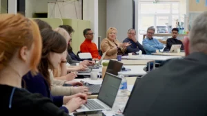 Vue en perspective d'une réunion professionnelle en groupe autour de tables modulaires, avec plusieurs participants utilisant des ordinateurs portables et une femme prenant la parole au centre.