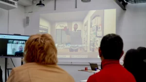 Vue de dos de participants à une réunion regardant une projection murale d'une simulation virtuelle sous-titrée et un écran de visioconférence sur le côté.