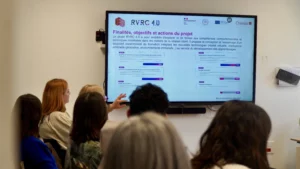 Vue de dos d'un public attentif devant un grand écran de télévision affichant les finalités et les objectifs du projet RVRC 4.0.