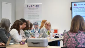 Profil d'une rangée de participants attentifs assis à une table de réunion, écoutant une intervenante au centre de la salle.